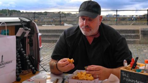 A man in black clothes and a black cap sits at a table outside eating fish and chips. A phone attacked to a tripod is on the table beside him. 
