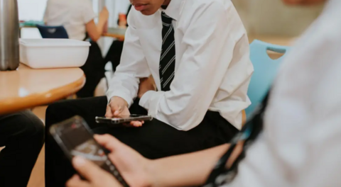 A boy in a shirt and tie on a phone sitting in a classroom.