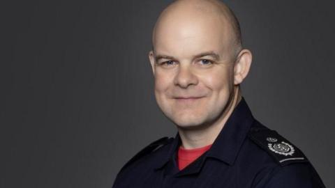 Formal work photo of Jonathan Smith, a bald man wearing a red T-shirt under a navy fire brigade boiler suit. There is a plain background and he is smiling.