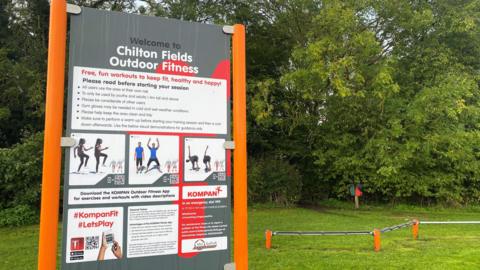 A grey sign attached to orange posts which reads Welcome to Chilton Fields Outdoor Fitness. It is in a field with trees in the background. 