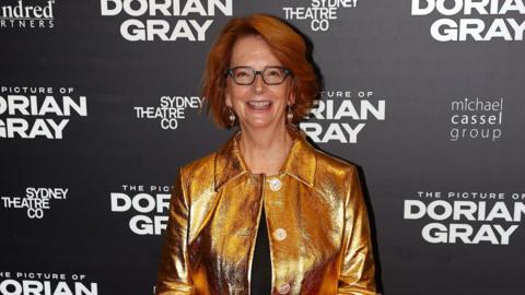 Julia Gillard attending an award ceremony. She has black glasses on and is wearing a gold jacket over a black top. Behind her is a black backdrop with the words The Picture of Dorian Gray in white, along with Sydney Theatre Co and Michael Cassel group. 
