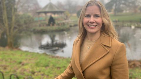 A woman wearing a tan coat and beige jumper stands in a park, smiling at the camera.
