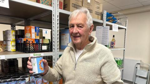 A man with grey hair and a pale hoodie holding a can of beans in one hand and standing in front of shelves containing more cans