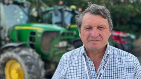 Robin Milton is standing in front of a green tractor. He is a wearing a checked shirt and a lanyard. There is another farm vehicle in the background.