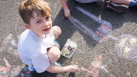 A little boy in a white polo shirt looks up at the camera as he chalks on the road