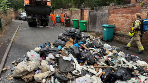 A pile of charred rubbish blocking the road, with a firefighter nearby, and a bin lorry parked up