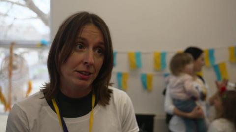 A woman with shoulder length brown hair. She is looking to the right. She is wearing a black vest with a white t-shirt on top and a black and yellow lanyard. In the background is a woman holding a child and a another person looking at her. Behind them is a white wall with bunting of the Ukrainian flag. 