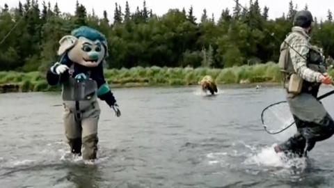 Someone dressed as Buoy the NHL mascot and wearing high waders, is seen running in knee deep water while being chased by a bear. Another man wearing fishing gear is also fleeing the running animal. In the background is a forest. 
