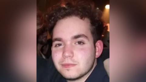 Head and shoulders photograph of Connor. He has dark wavy hair and is standing in a dark room with a spotlight in the background.