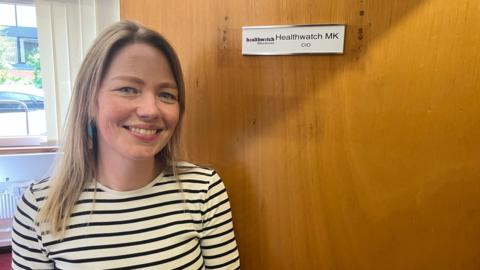 Maxine Taffetani with dark blonde hair down to her shoulders, who is wearing a white top with black horizontal stripes. She is stood by a door which has a logo for Healthwatch MK on it.