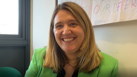 Georgia Gould is sitting down and is facing the camera, but looking towards the left of the frame. She is wearing a green suit jacket and is smiling.