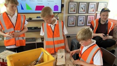Three boys, in orange high vis jackets building bridges with lolly stick inside the signal box