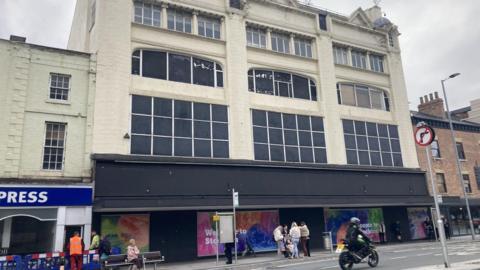 The weathered shop-front of a former Debenhams store. It has multicoloured boards reading Welcome to Stockton along the ground floor. The boarded up windows higher up the building are worn.