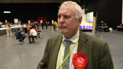 George Duggins stands in the count hall and wears a green suit jacket, white shirt, green tie and a red Labour rosette.