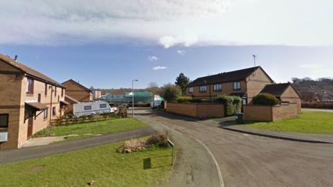 A cul-de-sac with terraced houses opposite one another. There is a white caravan parked on a driveway. There are grassy front lawns and trees in the background. There are blue skies and white cloud.