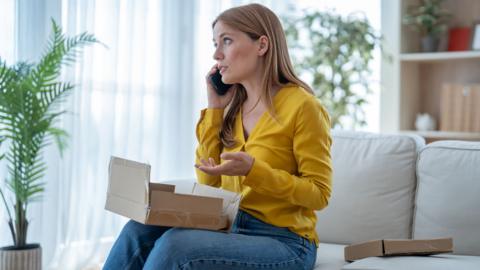 woman sitting on sofa at home, holding damaged cardboard box while complaining on phone