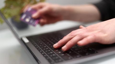 A close up of someone's hands with the left hand on the keys of a laptop and the right hand holding a bank card.