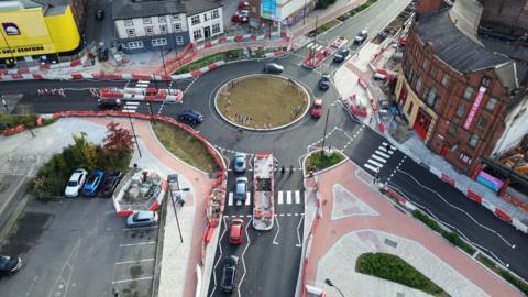 An image from a drone of a large roundabout, viewed from a height, with four exits surrounded by orange fencing. Some cars and vans are driving around it.