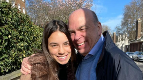 Hannah and Mike Lynch hugging and smiling into the camera. Miss Lynch has long brown hair and a brown coat. Mr Lynch has a blue jumper and shirt collar, and a black coat.