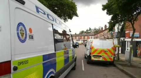 Two West Midlands Police vans are parked either side of a road, with trees either side. There are shops and terraced houses in the distance.