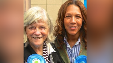 Two women smile at the camera. The one on the left has grey bobbed hair and the one on the right has shoulder length brown hair. Both are wearing blue rosettes.