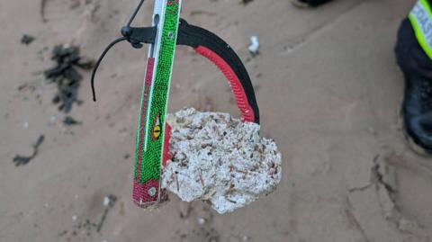 A man using a litter picker holds a white, waxy lump while standing on a sandy beach