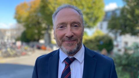 A picture of Paul Montague. He has grey hair and a grey beard. He is wearing a white shirt with a navy suit jacket and navy and red tie.