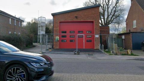 A small fire station with a red door and metal fencing outside it.