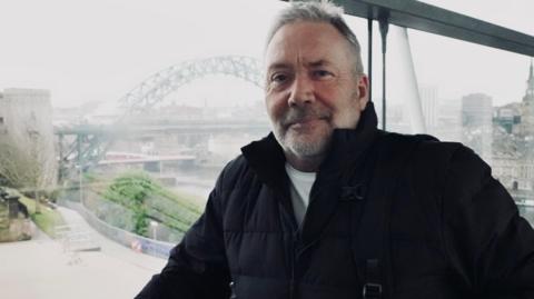 Andrew Parker is standing in front of a large window overlooking the Tyne Bridge. He is wearing a white t-shirt and a black puffer jacket. He has grey hair and beard and is carrying a backpack.