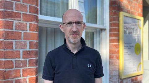 A bald man wearing glasses and a navy blue polo shirt. He is standing outside a railway station.