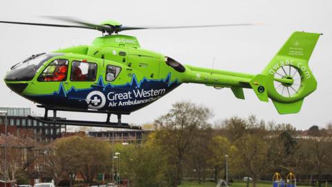 Green and blue helicopter with the word 'Great Western Air Ambulance' written across it. It's taking off over a green park area that is near a road. The sky is grey and cloudy. 