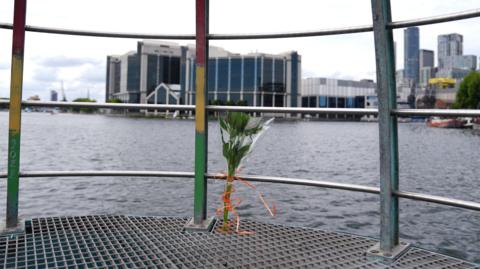 Bouquet of flowers tied to a metal railing overlooking a river, with modern buildings in the background.