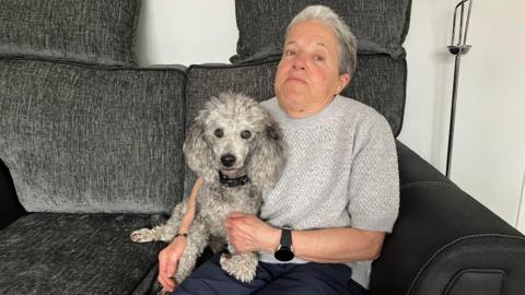 An woman sits on a dark grey sofa holding a small curly-haired dog on her lap. The dog looks directly at the camera while wearing a collar, with soft textured fur visible.