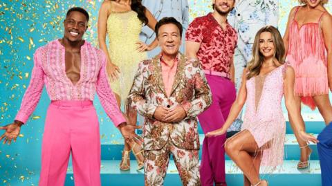 Three ballroom dancers, a man in a pink jumpsuit, a man in a patterned suit and a woman in a pink sparkly dress.