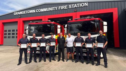 Firefighters in uniform holding certificates, in front of two fire engines.