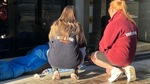 Welcome Organisation workers kneel on the ground in an attempt to speak to a person in a blue sleeping bag