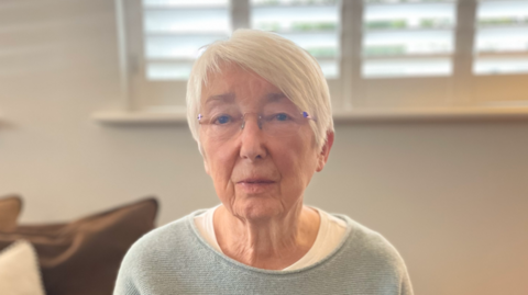 A woman in a blue jumper. Elderly, wearing frameless glasses. She has grey/white short hair and sits inside a living room.