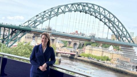 Rachel Reeves, who has dark brown hair, is wearing a navy blue suit and is standing on a balcony, leaning against a railing. She is smiling and facing the camera. Behind her is the green Tyne Bridge. It is almost fully visible with part of both the High Level Bridge and Swing Bridge in view behind it. Reeves stands on the Gateshead side of the river and part of Newcastle Quayside can also be seen.