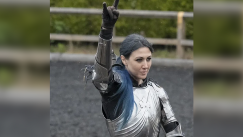 A woman with long blue hair wearing a breast plate makes a bull horns hand gesture.
