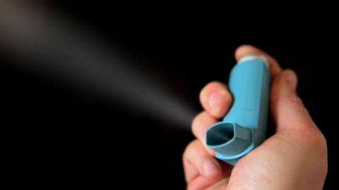 A close-up of a hand holding a blue inhaler, against a black background.
