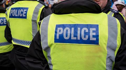 Stock photo of a police man facing away form the camera. He is wearing a black jacket with a yellow high vis jacket that reads POLICE on the back.