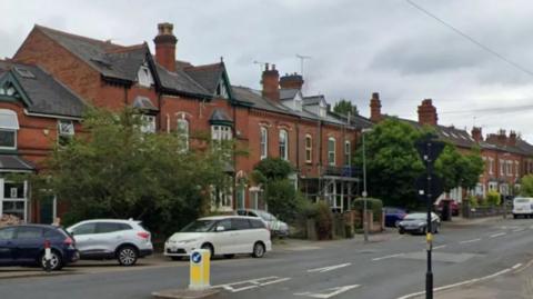 A residential street with cars parked in front of a row of red brick houses and a traffic bollard in the middle of the road