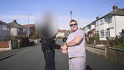 Police dashboard camera footage showing James O'Donnell, 35, who is wearing a lavender t-shirt and matching shorts and black sunglasses. He is standing in the middle of a residential road with a police officer in black uniform, who has their face blurred, placing him in handcuffs. O'Donnell is grinning towards the camera. 