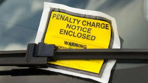 A yellow parking ticket warning of an enclosed penalty notice tucked under the windscreen wiper of a car.