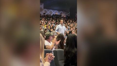 Man in a light blue shirt walking down the steps of an auditorium as the audience applause.