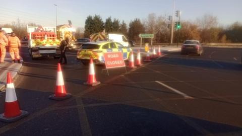 Red cones and road closure signs block the entry to a road junction where emergency vehicles are also parked.