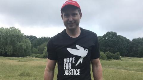 Aaron Robinson, aged 40, smiles at the camera wearing a black t-shirt with the Hope for Justice's charity branding. Behind him is a grassy field where he runs.