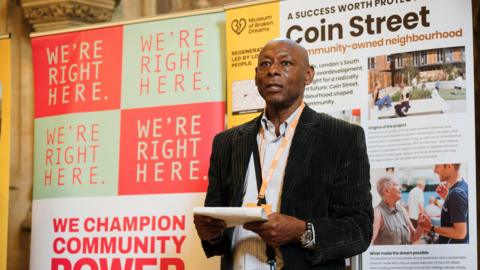 Image shows a man wearing a black suit delivering a speech. 
He is standing in front of two banners about the 'Museum of broken dreams' and the story of the Chapeltown Frontline Self-Build. 