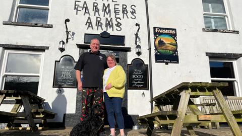 A man wearing a black top and patterned trousers is standing next to a woman with a yellow cardigan, white shirt and blue jeans.  
Sitting in front of  them is a black spoodle dog.
They are all in front of a white building with a sign saying "Farmers Arms" and there are wooden picnic benches either side. 