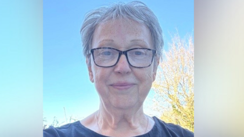 Christine Grant is seen from her shoulders up. She is wearing a dark blue T-Shirt and has short, grey hair. The sky behind her is blue.